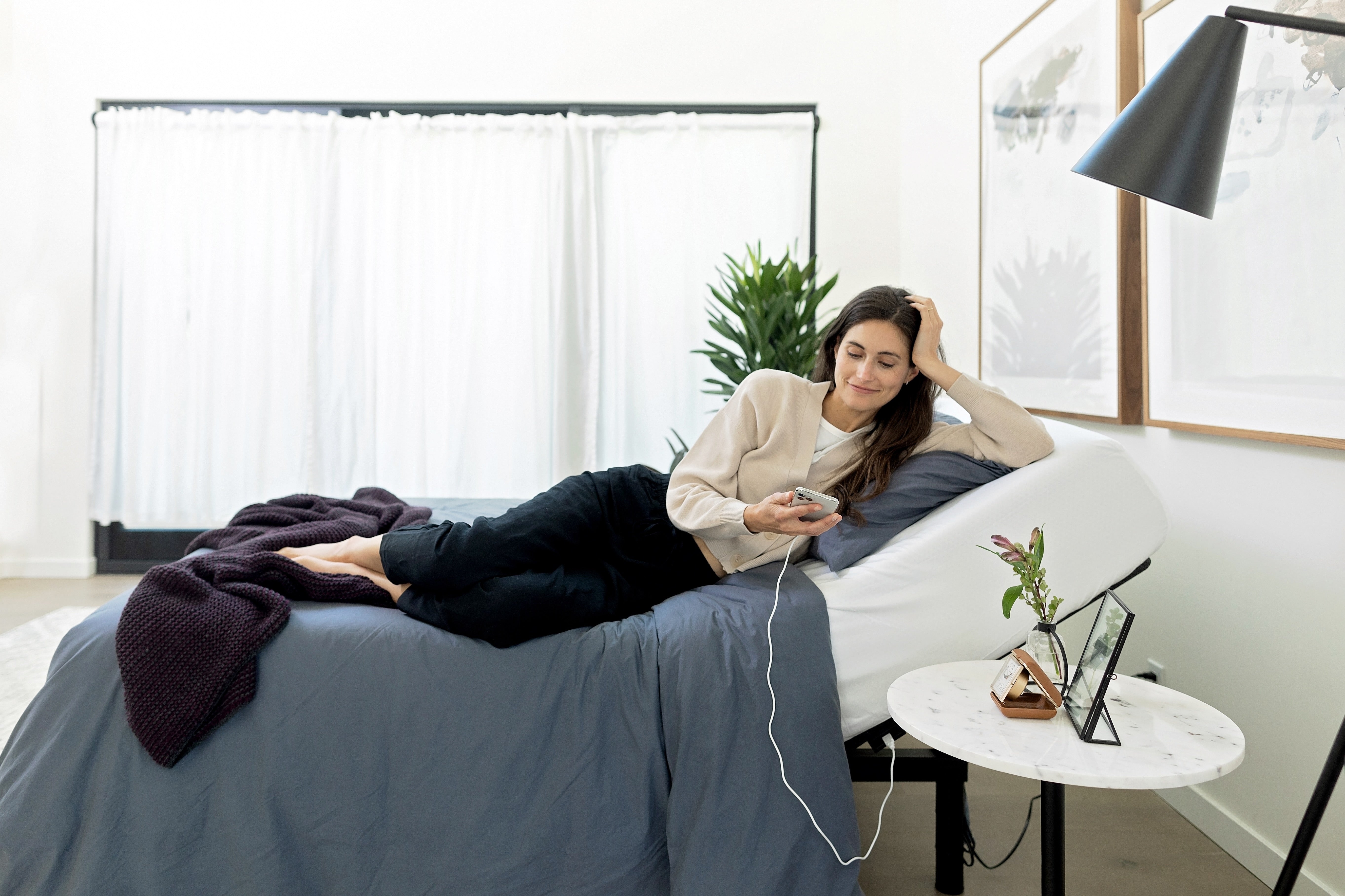 Woman lying on a bed using a smartphone as an adjustable base remote in a bright room with a plant and table.