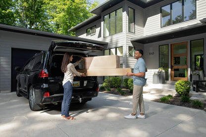 Two people unlading adjustable bed frame boxes from a black SUV in front of a modern house.
