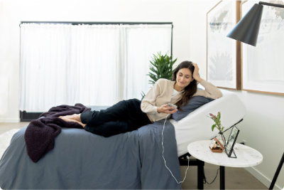 A person relaxing on an adjustable bed base in a bedroom setting. The bed is dressed in gray bedding, and there is a nightstand with a lamp and a plant beside it.