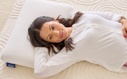 Woman lying on a bed with her head on a Ziwi pillow, wearing a white shirt.