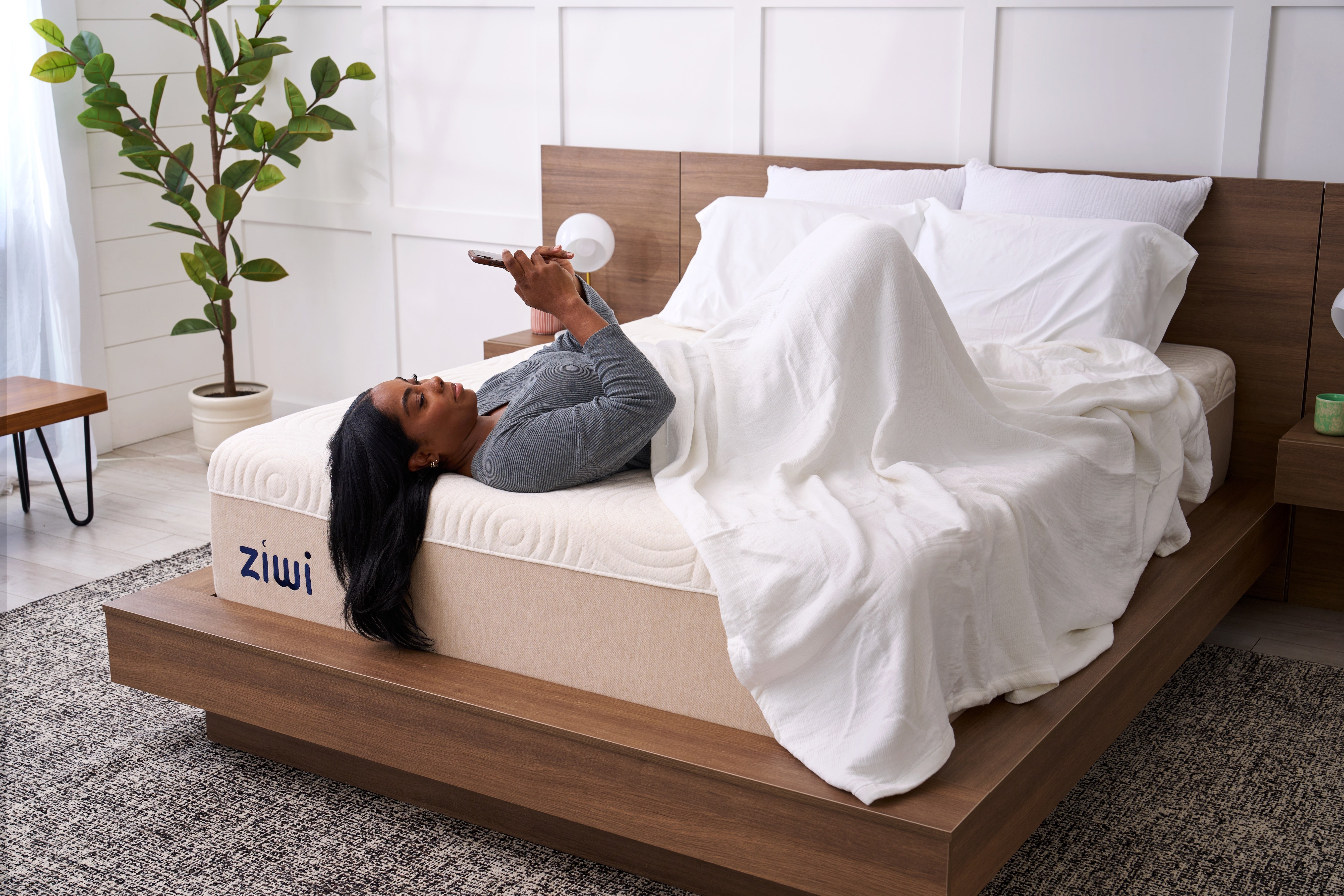 Woman lying on a bed with a Ziwi mattress, surrounded by a plant and a small table.
